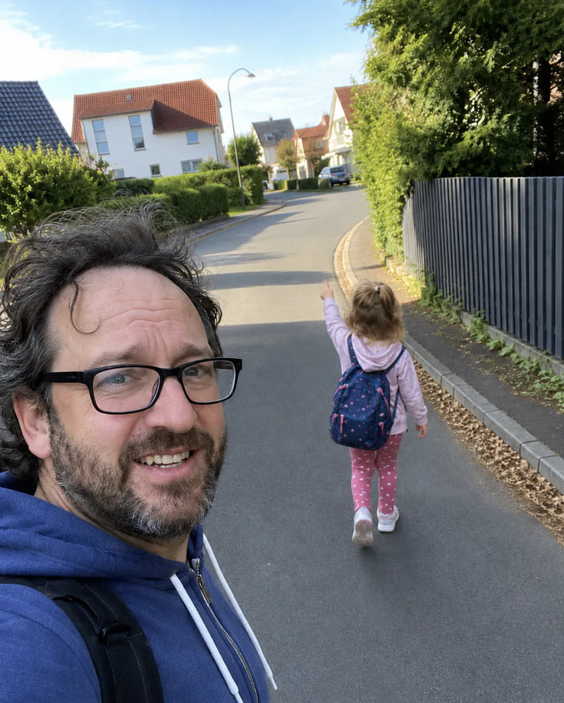 Smiling dad in glasses takes a selfie while his daughter in a backpack walks ahead down the street