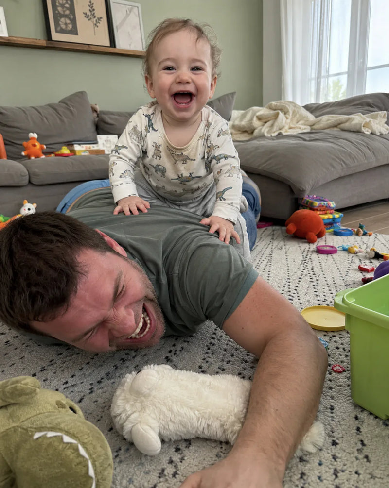 Giggling baby sits on dad's back while he lies face-down on the living room rug beside a stuffed toy
