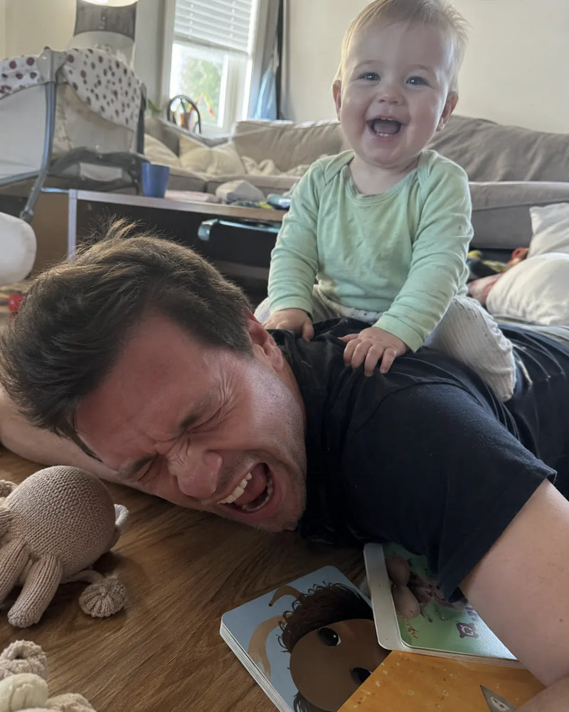Baby in a green sweater sits on dad's back while he winces on the floor next to picture books