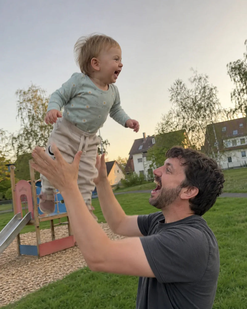 Dad lifts his laughing toddler high into the air on a grassy playground at dusk