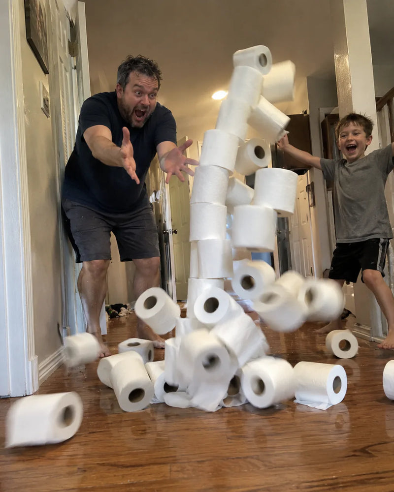 Dad dives to catch a toppling tower of toilet paper rolls in the hallway while his son cheers beside it