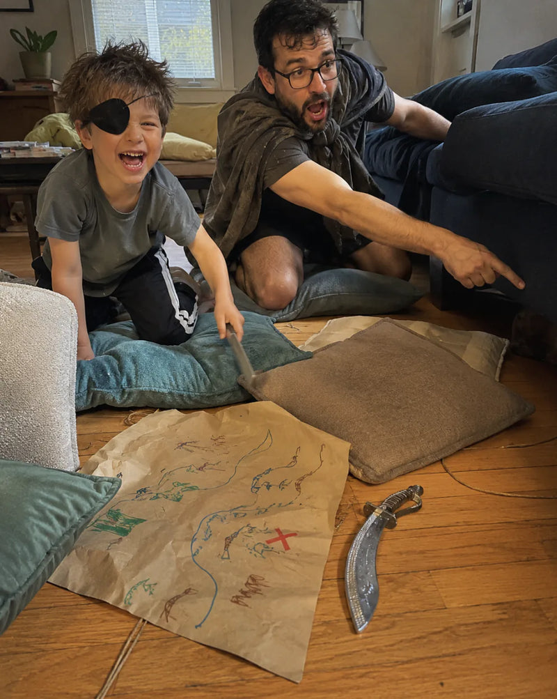 Boy in a pirate eyepatch and his dad play pirates on the living room floor next to a hand-drawn treasure map