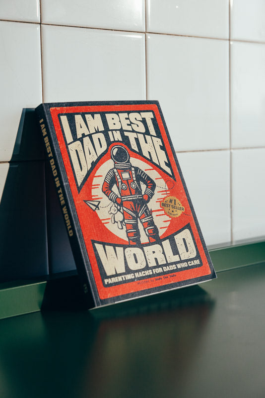 Red I Am Best Dad In The World book with an astronaut cover standing against white tiles on a green shelf