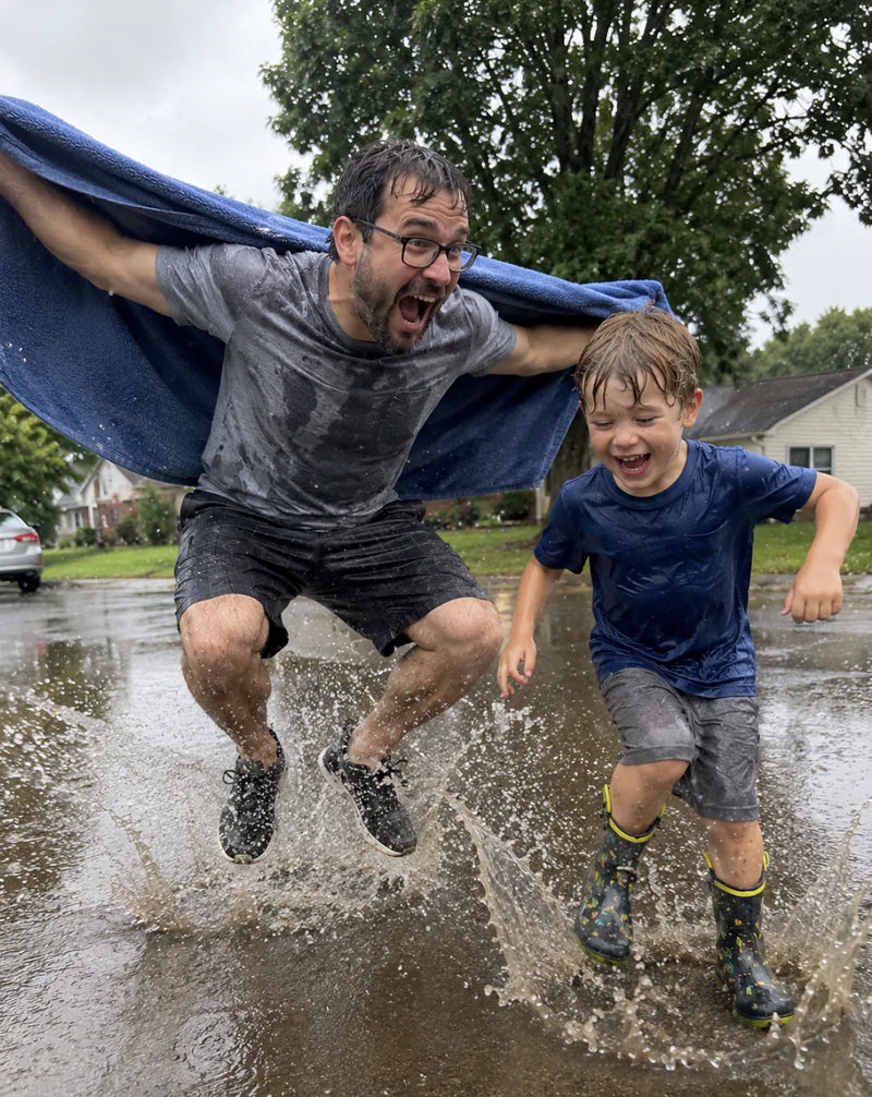 Dad and his young son jump through a rain puddle, a blue blanket spread behind dad like a cape