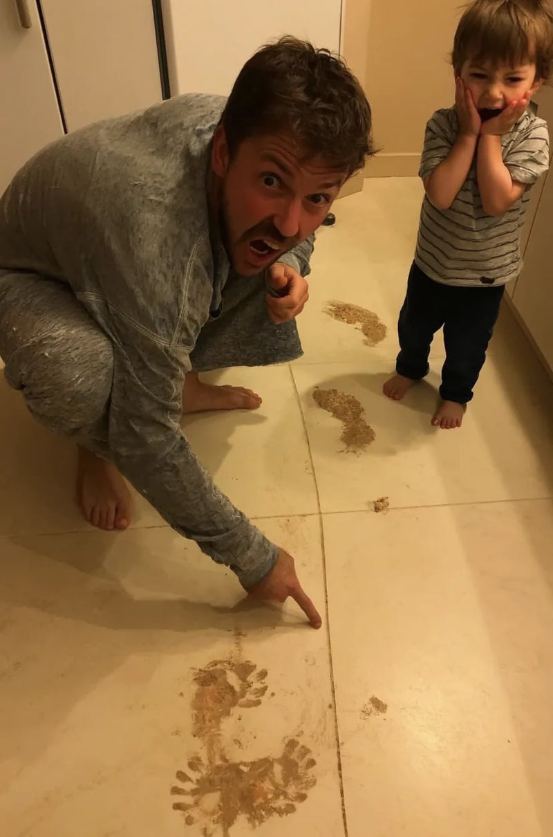 Dad crouches and points at muddy footprints across a tile floor while his son covers his mouth in surprise