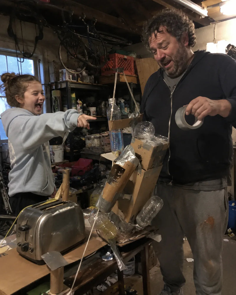 Dad in a cluttered garage workshop holds a roll of tape next to a lopsided cardboard robot while his kid points and laughs