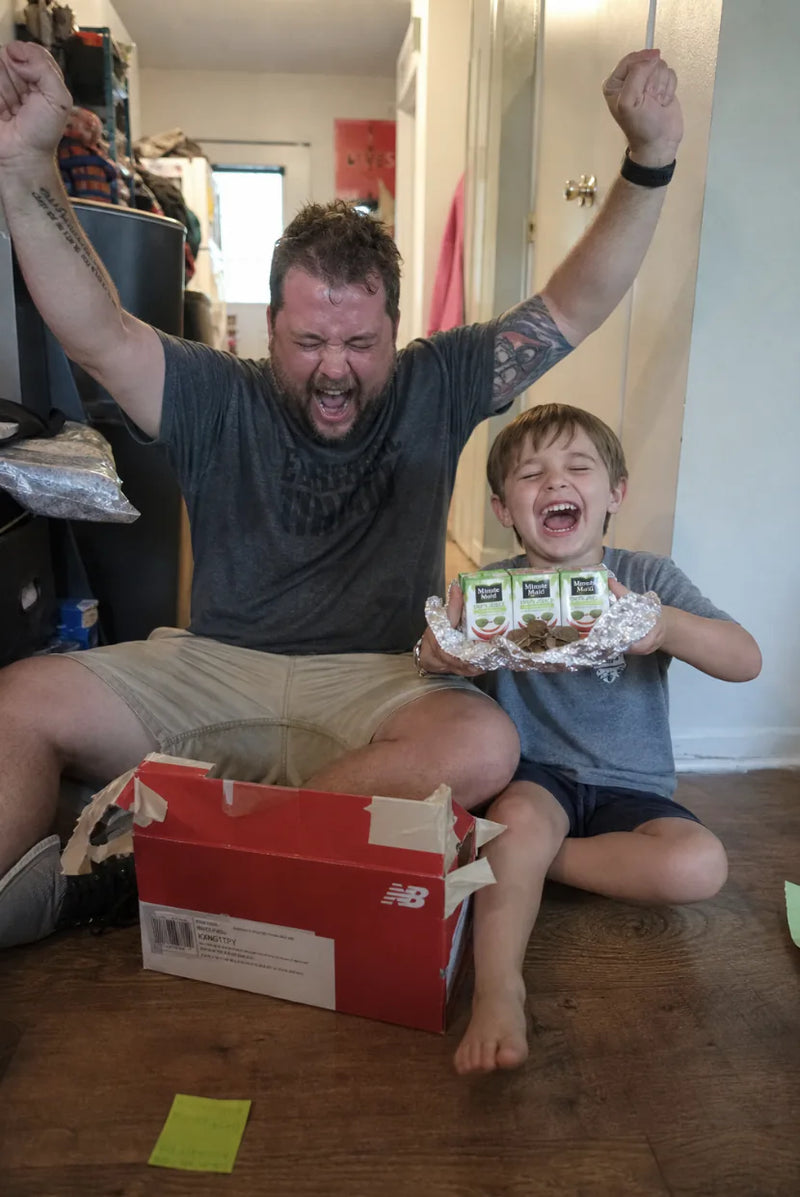 Dad cheers on the floor with a fist in the air beside an open shoebox while his son laughs, holding up a small cardboard model