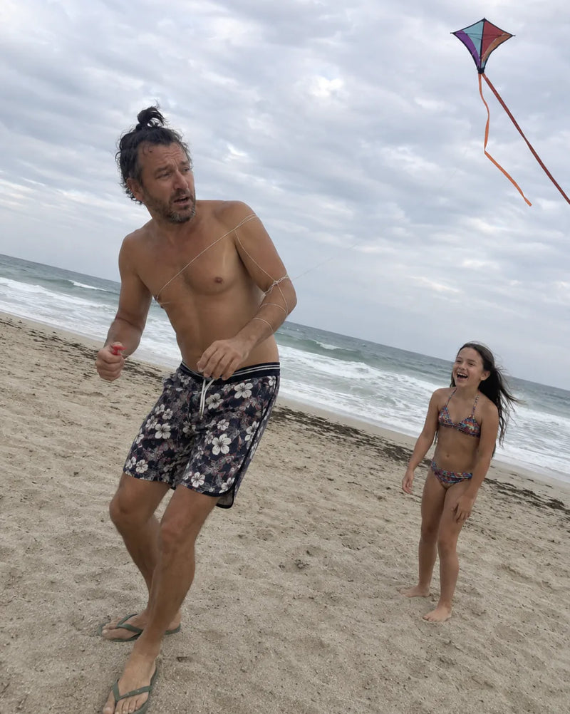 Dad in swim trunks sprints along a beach under a kite while his daughter watches from behind