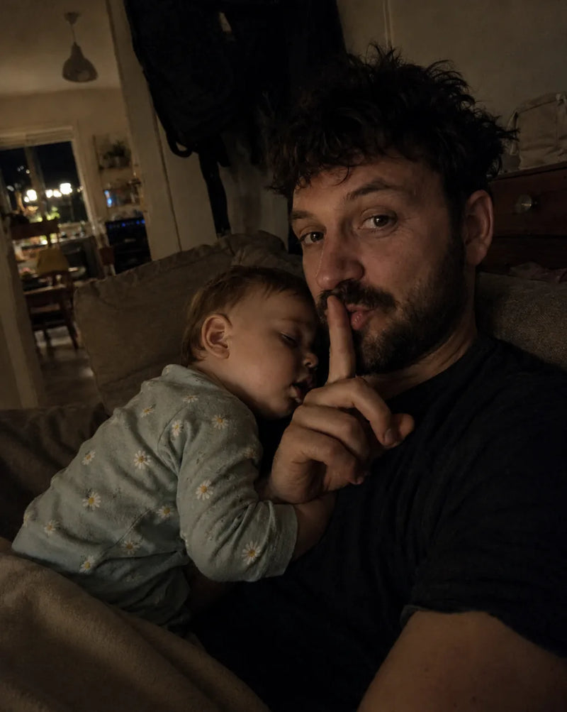 Dad holds a finger to his lips for quiet while his baby sleeps on his shoulder in the dim living room
