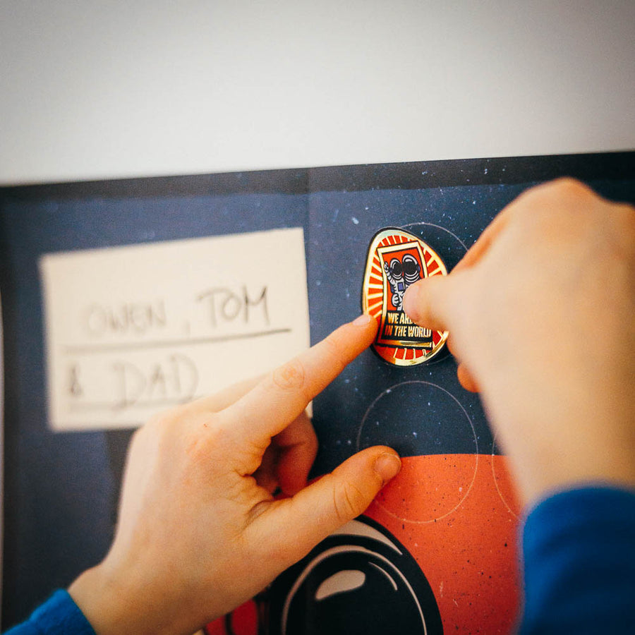Dad's hand presses a gold mission sticker onto the poster beside a handwritten name card reading Owen, Tom & Dad
