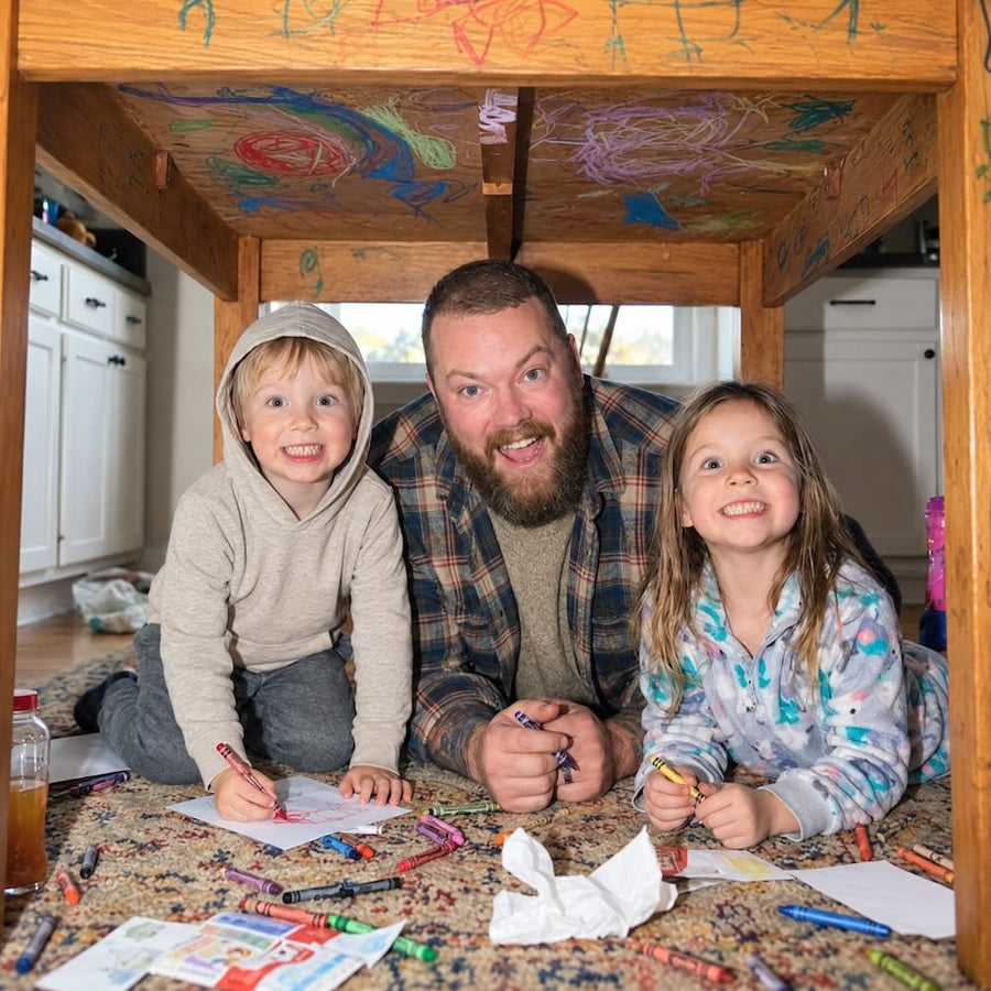 Man with two children sitting under a wooden loft bed with colorful drawings on the wall.